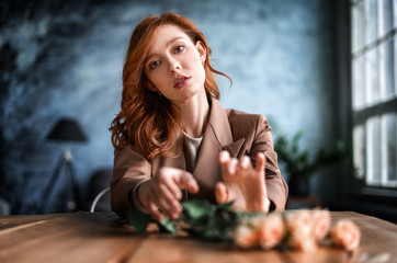 Portrait of a pretty red-haired girl with flowers sitting at the table. The girl is dressed in a brown jacket.