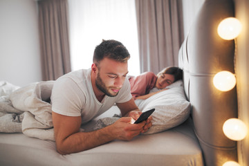 Man using phone in bed while woman sleeping.