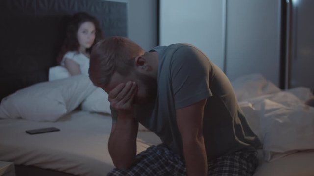 Medium shot of exhausted young Caucasian man sitting on side of bed, with smartphone beside pillow, and rubbing forehead, and angry woman glaring at his back