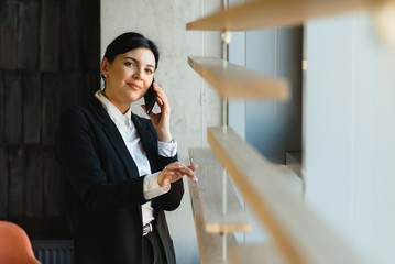 Cheerful young business woman standing in modern office talking on her mobile phone. Beautiful female model using cell phone.