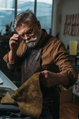 attentive shoemaker touching glasses while looking at piece of genuine leather