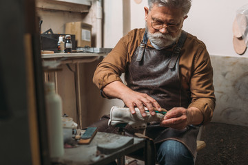 Selective focus of senior, bearded shoemaker repairing shoe in workshop