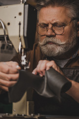 selective focus of attentive, senior shoemaker sewing leather on sewing machine