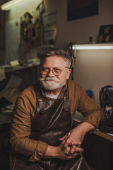 smiling, senior cobbler looking at camera while sitting in workshop