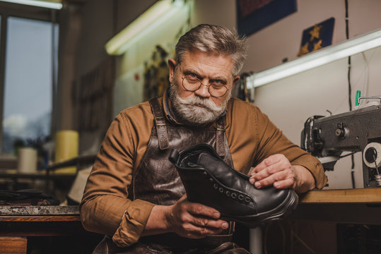 serious, senior shoemaker holding leather boot while looking at camera - Powered by Adobe
