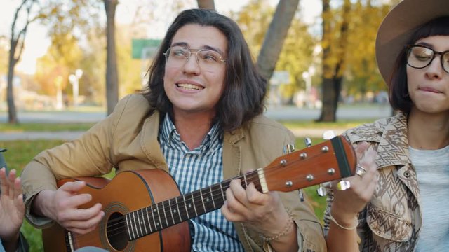 Portrait of creative youth girls and guys singing playing the guitar relaxing in urban park together. Leisure time activities and modern lifestyle concept.
