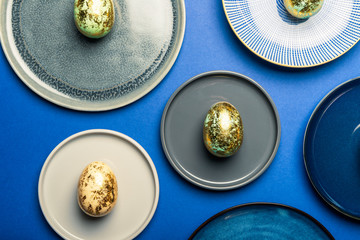 Different Ceramic Plates with Golden Speckled Easter Eggs of pastel colors on blue background. Happy Easter card concept, minimalistic design, top view