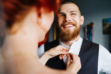 Beautiful bride and groom together in the bright room before the marriage ceremony. She's buttoning his suit waistcoat. Man giggles, feeling a little anxious.
