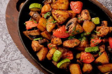 Delicious oven-baked potatoes with herbs and vegetables in a pan, rustic style. Potatoes fried, red cherry tomato, zucchini, onion on gray stone background.