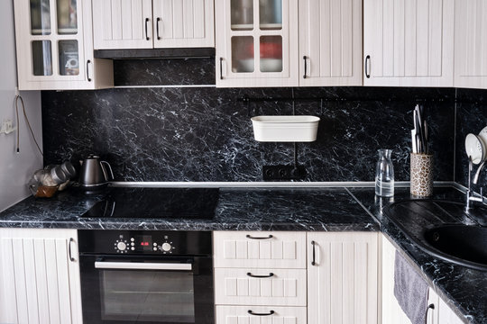 Modern kitchen is white with a black marble countertop. Stove, oven, cabinets and sink with tap