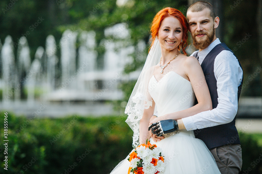 Wall mural Portrait of the handsome newly wed couple standing together in the park in front of a fountain. Man hugging his wife from behind. He has a prosthetic bionic arm.