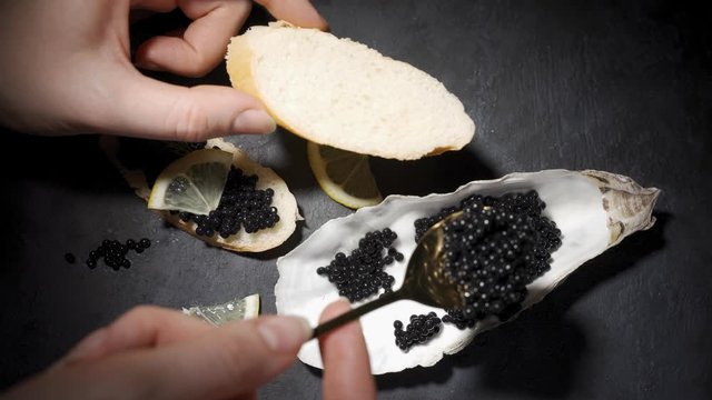 Woman smears black caviar on a baguette. Caviar in a oyster shell on a black background
