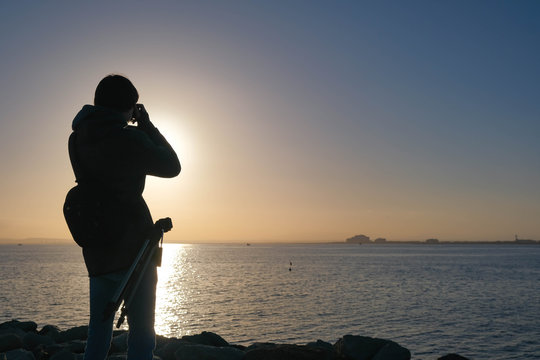 Young photographer takes photos the sunset over the Bay of Roses, Catalonia, Spain. Traveling with a camera along the European coast. Man sets up the camera for shooting.