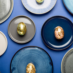 Different Ceramic Plates with Golden Speckled Easter Eggs of pastel colors on blue background. Happy Easter card concept, minimalistic design, top view