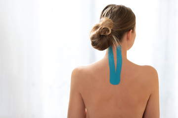 Female patient with kinesio tape on her upper back and neck standing against white background, rear view. Kinesiology, physical therapy, rehabilitation concept.