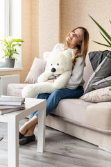 Cute young girl in a white sweater and jeans with a lovely smile sits on a sofa with a white teddy bear in her arms
