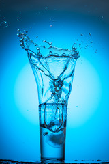 large splash and stream in a glass with clear water on a blue cyan background from an ice cube