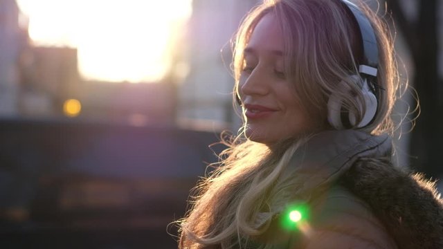 Happy woman listening to music on wireless headphones connected to mobile phone, on the streets at sunset, slow motion