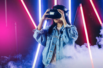 Mod curly dark haired girl dressed in blue denim jacket uses the virtual reality glasses on her...