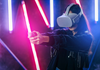 Mod curly dark haired girl dressed in blue denim jacket uses the virtual reality glasses on her head in the dark studio with neon light and smoke fog with place for your text