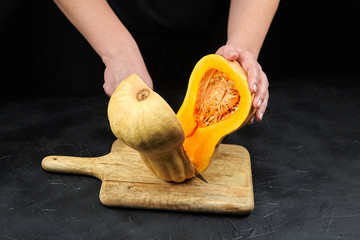 Butternut squash halves on wooden chopping board on black background. Female hands with steel...