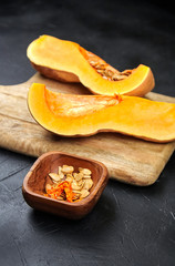 Pumpkin seeds in wooden bowl, Butternut squash pieces, cutting board on black background, closeup. Cooking winter squash