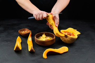 Butternut squash, woman cuts pumpkin peel. Female hands with steel knife, wooden bowl on black background. Cooking sweet winter squash