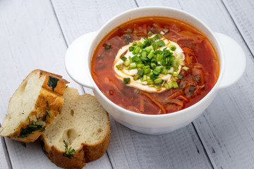Ukrainian red borscht with sour cream in white bowl and pieces of bread with garlic. Russian cuisine concept. Light wooden planks background, table.
