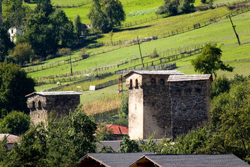 Fototapeta premium Svan towers, stone, quadrangular, against the background of green pastures and meadows, fenced off by fences in a mountain valley on a bright sunny day. Mestia Village, Svaneti, Georgia