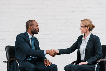 side view of happy african american businessman shaking hands with businesswoman while sitting on chair