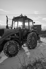 Fototapeta na wymiar Agricultural tractor with a trailer rides along a country road covered with snow in the early winter morning, in the sun. Black-and-white photo.