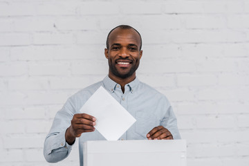 happy african american voter putting blank ballot in voting box