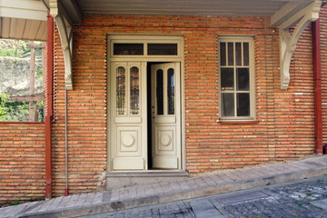 Old wooden white door in a brick house. The door is ajar. Near the same old window. A road passes under a very strong slope past the house. Old Town, Tbilisi, Georgia