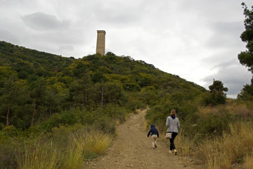 Svan Tower on the top of a hill in the Tbilisi Ethnographic Museum. A woman and a child walk along a narrow, overgrown path leading through the trees to the tower. The sky is gloomy and gray