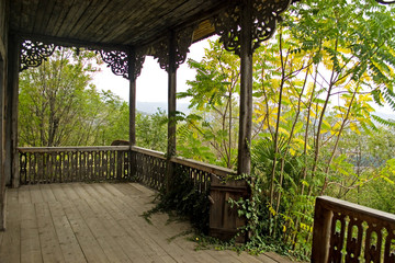 Empty abandoned wooden terrace of an old Georgian house. A wild forest surrounds the terrace and penetrates it to a balustrade. Columns decorated with carved wood ornaments