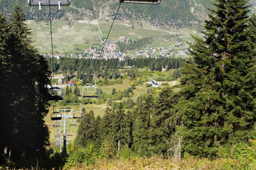 The slope of the ski resort in the late summer. Empty chairs of the funicular. Around green spruce. In the background on the mountainside you can see the Georgian village of Mestia