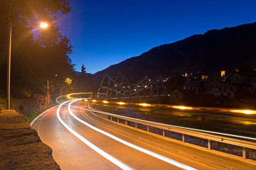Twilight on the mountain road. Long exposure shot. The lights of the car with flasher in the form of glowing tracks. Against the background of the bridge, mountains, village. Mestia, Georgia