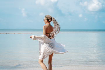 very slender blonde with dreadlocks against a tropical landscape with a blue sea and sky and, in a white shirt and sunglasses, tropical vacation and travel concept