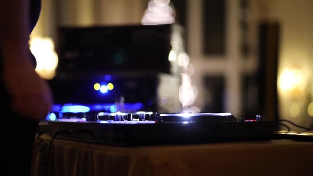 DJ uses scratch performance DJ music controller mixing console at nightclub party venue. Party lights and blurry background