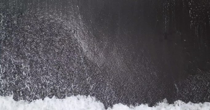 Top down aerial view of waves on black sand beach in Amed, Bali, Indonesia. Drone moves up and reveals the beach.