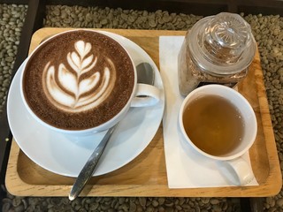 The hot coffee that there is latte art on top. There is a cup of tea beside coffee cup on tissue paper. Every things are on wooden tray. Background is coffee nut.