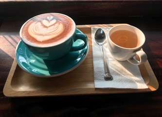 The cup of coffee that there is heart latte art on top.  There is a cup of tea beside coffee cup on tissue paper. Every things are on wooden tray. There is soft sunshine to them.