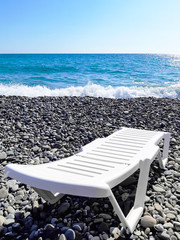 One white plastic sun bed stands headboard to the viewer on a pebble beach on the sea shore with small waves against the background of a clear blue sky. Vertical orientation, major plan.