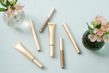 Mascaras in different gold packages on a blue background next to a vase of flowers. The concept of makeup, fashion and beauty