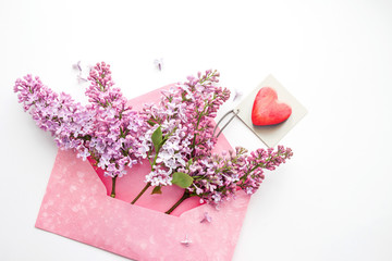 Red heart on a card and lilac flowers in a pink envelope on a white background, top view. Romantic card by a holiday	