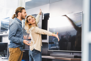 smiling girlfriend pointing with hands at tv and looking at shocked boyfriend in home appliance...