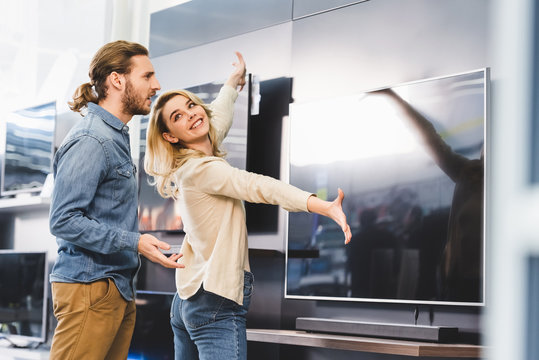 Smiling Girlfriend Pointing With Hands At Tv And Looking At Shocked Boyfriend In Home Appliance Store