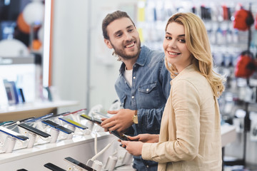 smiling boyfriend and girlfriend holding new smartphones in home appliance store
