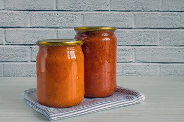 Two transparent glass jars with zucchini caviar and homemade adjika on a white wooden table against a brick wall