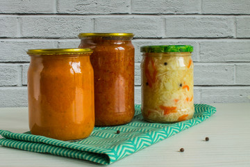 Three glass transparent jars with vegetable snacks: zucchini caviar, adjika and sauerkraut on a white wooden table against a brick wall